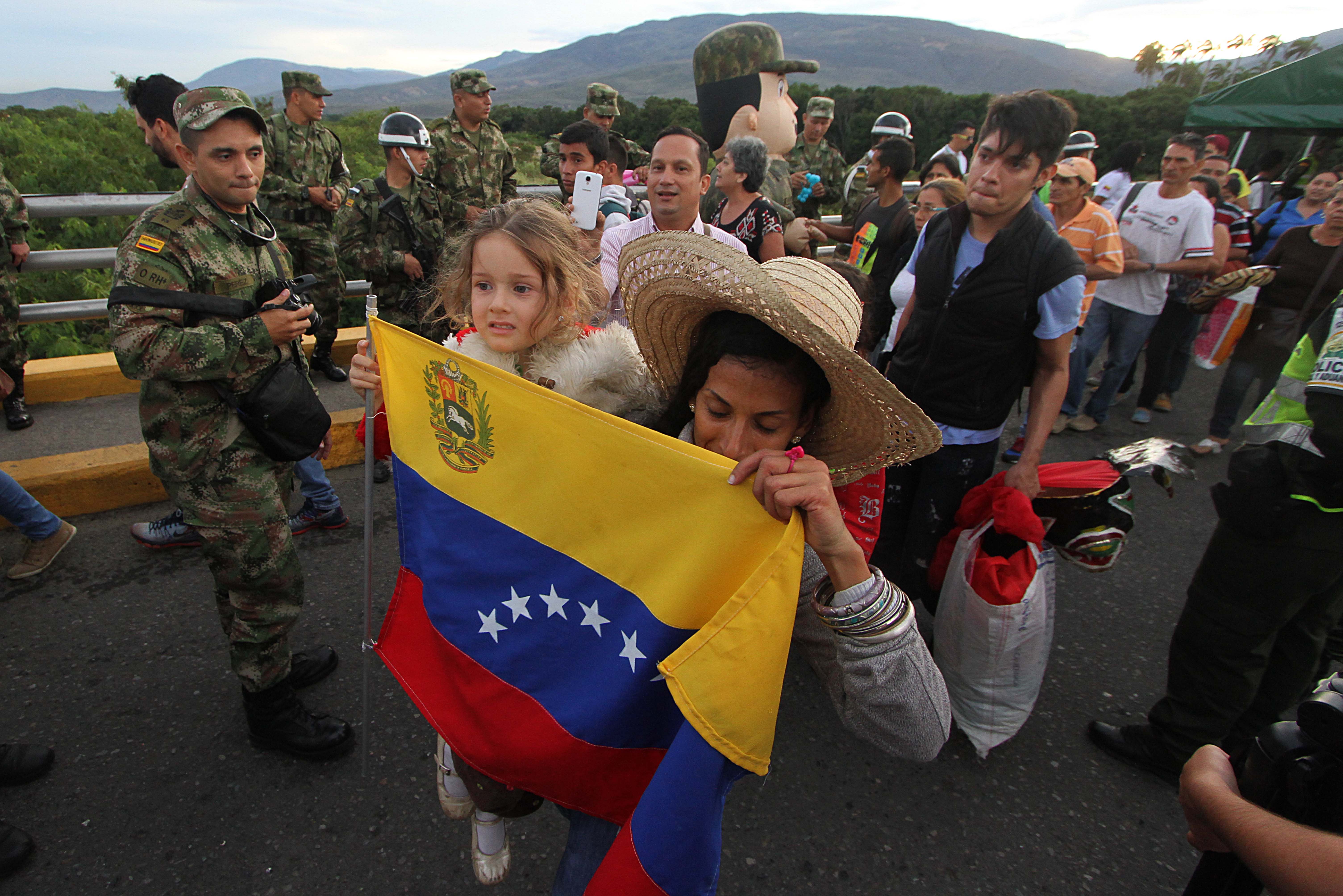 A Venezuelan refugee carrying a Venezuelan flag and her child cross the Simón Bolívar bridge between Venezuela and Colombia, past Colombian Army soldiers. J.R.S. photo by George Castellanos.