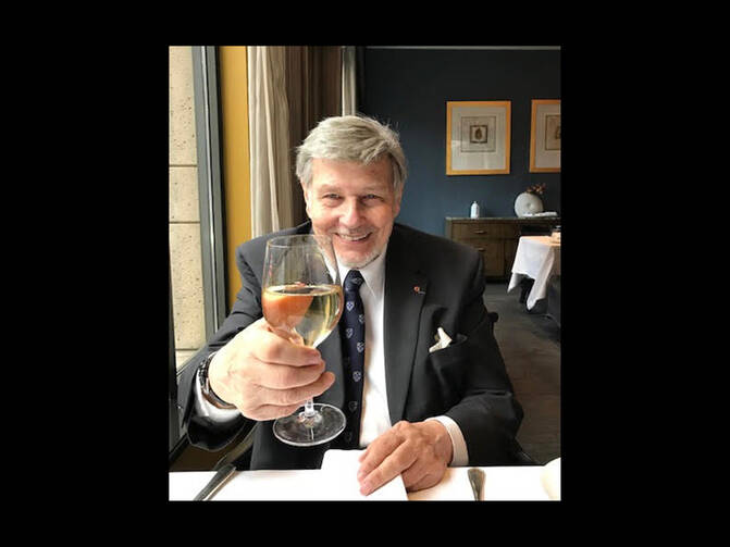Jesuit Jacques Monet sitting at a table in a restaurant, smiling and toasting with a glass of white wine. He is wearing a dark suit and a tie with a pin on his lapel.