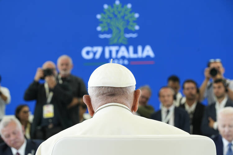 Pope Francis, seen from behind, sits at a working session during the G7 summit in Borgo Egnazia, Italy. The session discusses AI, Energy, Africa, and the Mideast. The background features a blue screen with the G7 Italia logo, and several attendees and photographers are visible.