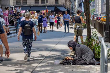 A mature homeless man sits next to a tree on the sidewalk of Fifth Avenue in Manhattan, reading a book while people walk past him. (iStock/carstenbrandt)